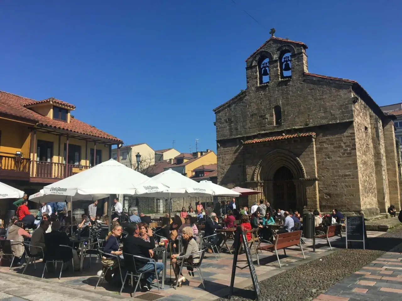 Gente disfrutando de un día soleado para comer en Avilés, con mesas al aire libre frente a una iglesia antigua.