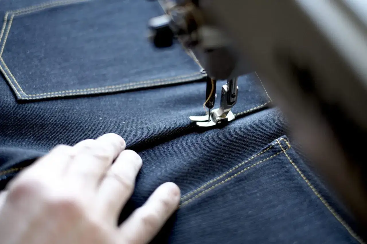 A hand guides dark denim under a sewing machine, creating a classic jeans template with yellow stitching.
