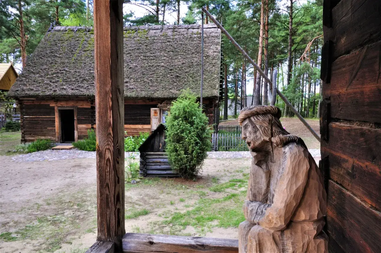 Drewniana rzeźba postaci, tradycyjna chata z mchem na dachu, zielone drzewka i płot. Wspaniałe **kadzidło atrakcje** skansenu.