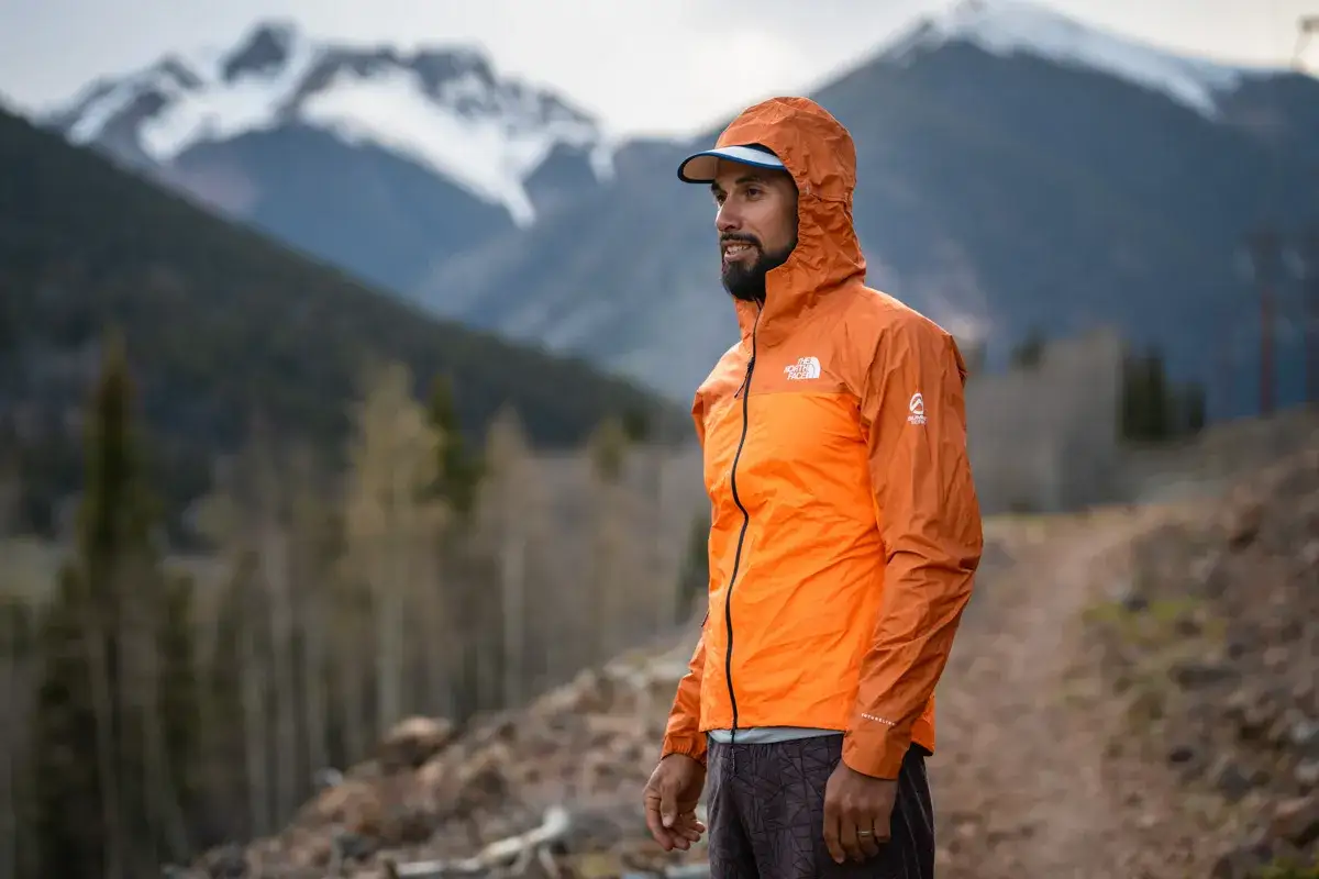 Mann in orangefarbener Jacke mit Kapuze, bereit für das Laufen bei Regen in den Bergen.