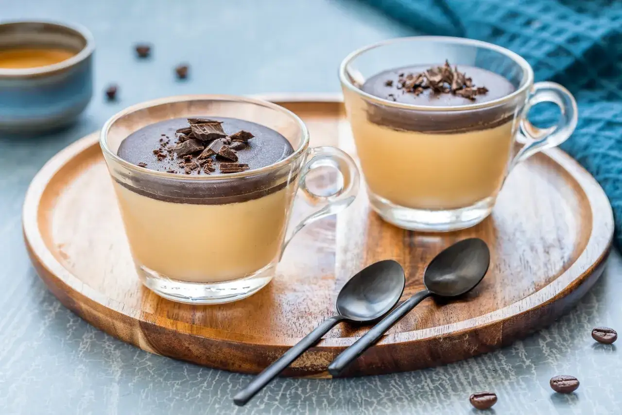 Due bicchierini di cremoso cremino al caff&egrave;, guarniti con cioccolato fondente e serviti su un vassoio di legno.