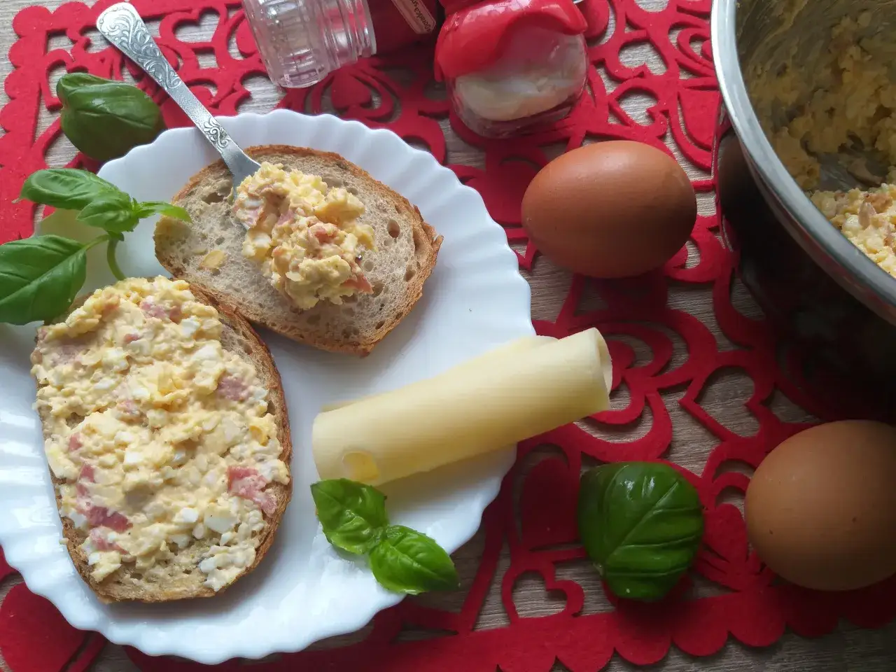 Dwie kromki chleba z obficie rozsmarowaną pastą jajeczną, obok ser, jajka i bazylia.