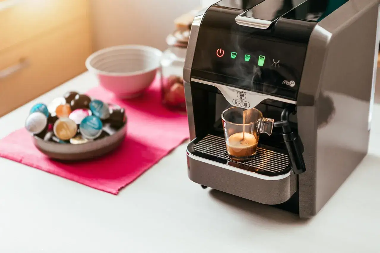 Macchina per il caff&egrave; che prepara un espresso fumante, con una ciotola piena di capsule colorate accanto.