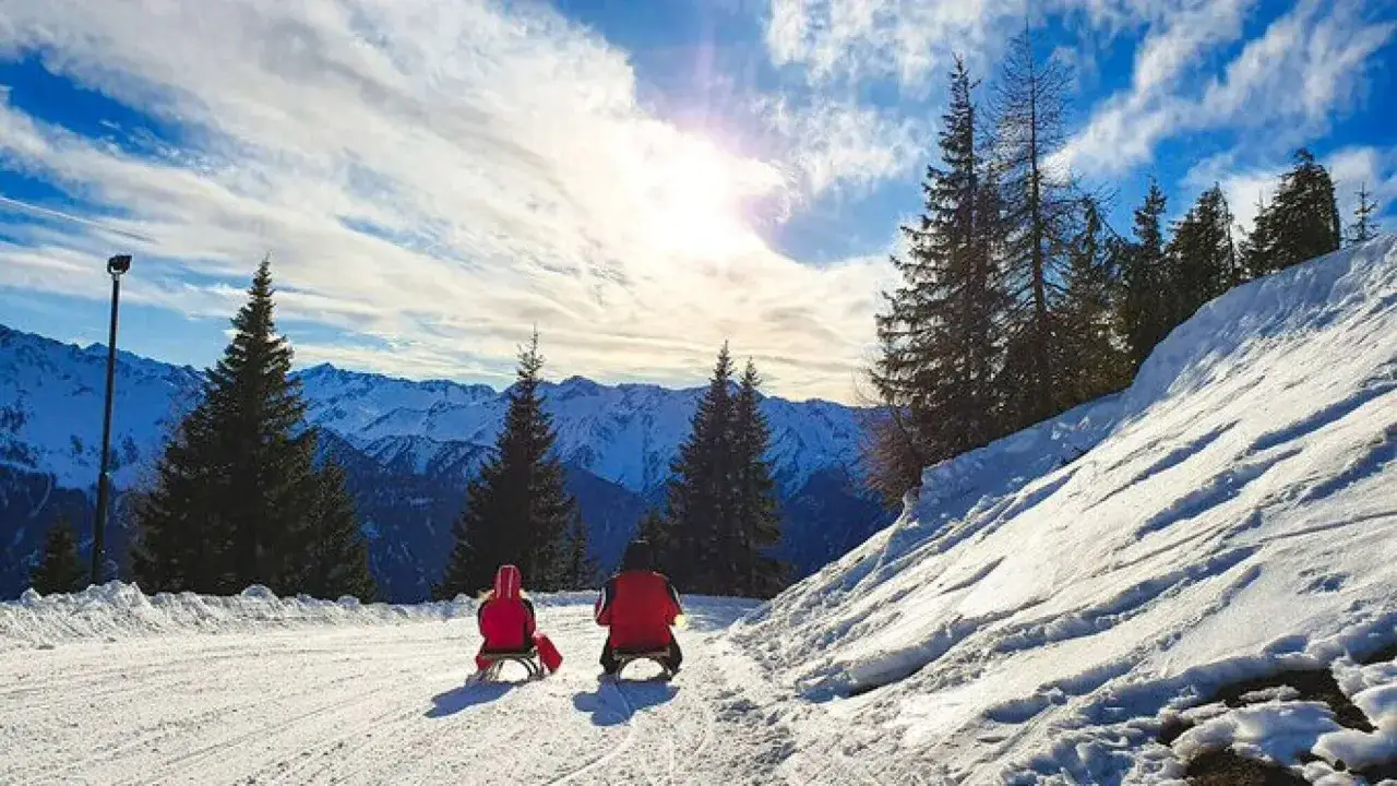 Zwei Personen rodeln auf einem verschneiten Hang in Bayern. Die Sonne scheint durch die Wolken auf die Berge.