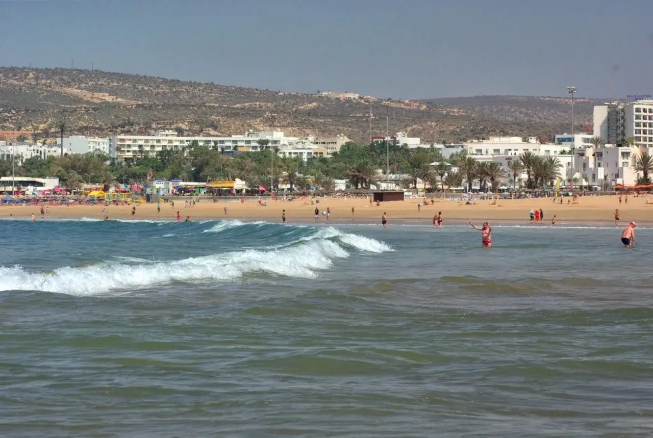 Agadir pogoda marzec: plaża pełna ludzi, palmy, białe budynki i wzgórza w tle. Fale na oceanie.