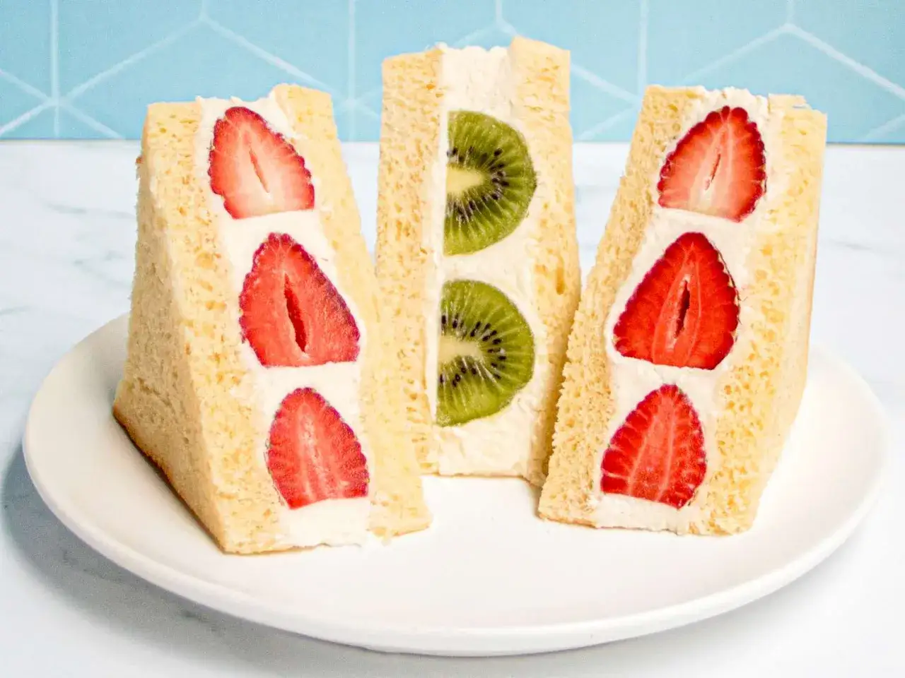 Three triangular Japanese sandwiches with fresh strawberries and kiwi slices nestled in whipped cream between soft bread slices.