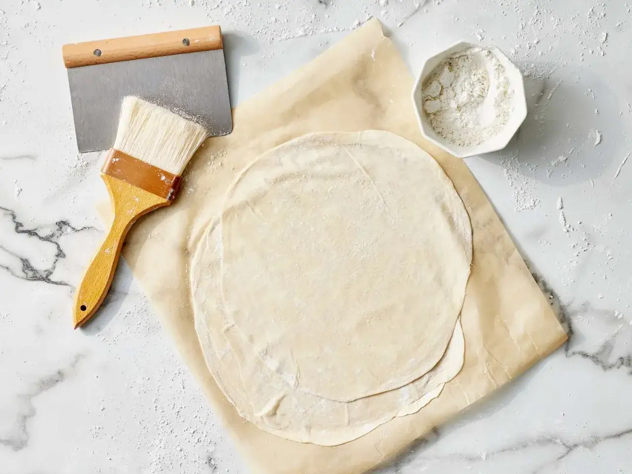 Preparando **que es la pasta filo**: finas láminas de masa, un pincel y un bol con harina sobre una superficie de mármol.