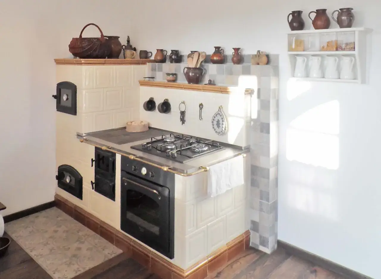Kuchenny piec kaflowy z płytą gazową i piekarnikiem, obudowany kremowymi kaflami, ozdobiony ceramicznymi dzbankami.