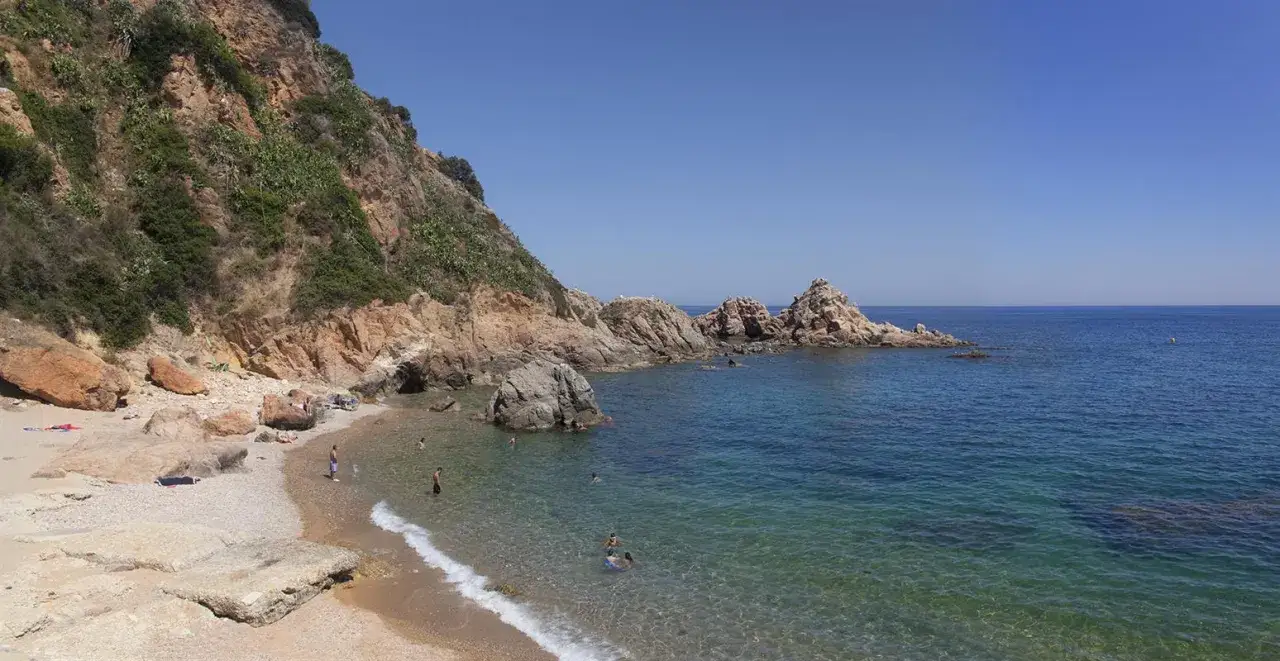 Les platges de Blanes ofereixen aig&uuml;es cristal&middot;lines i roques imponents sota un cel blau. Gent gaudeix del sol i del mar.