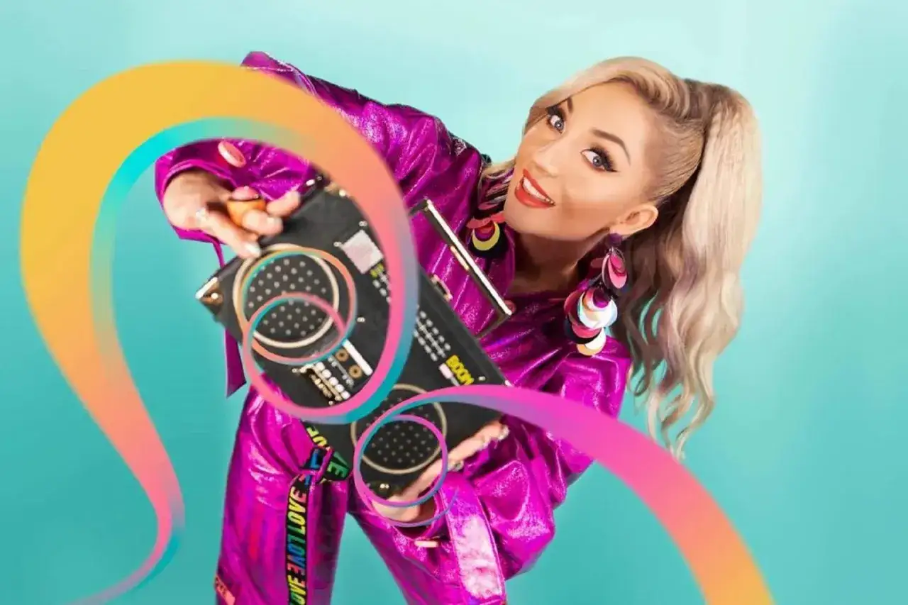 Cleo w błyszczącym, fioletowym stroju prezentuje nową piosenkę, trzymając w rękach retro boombox. Kolorowe ozdoby i tęczowy motyw podkreślają jej energię.