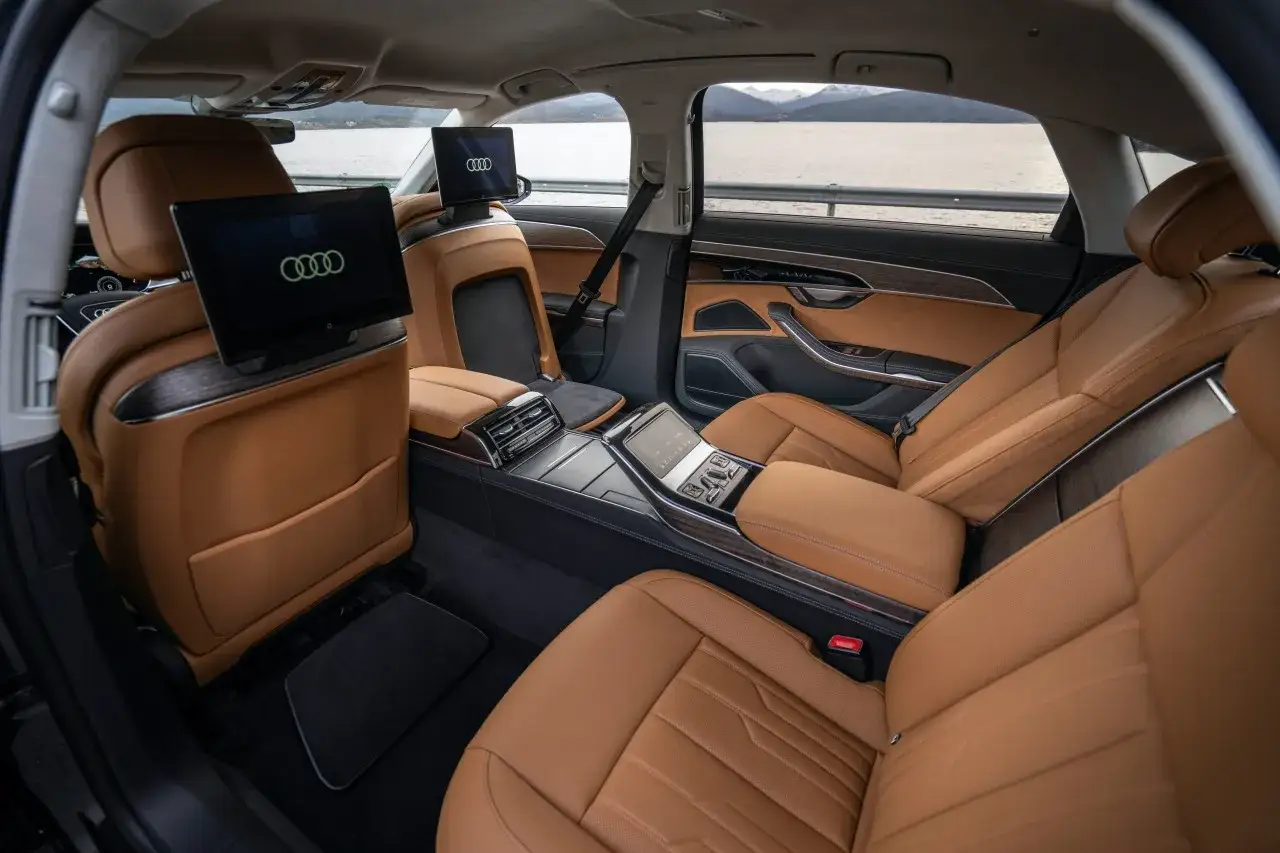Luksusowe wnętrze Audi A8 2025 z brązową skórzaną tapicerką, ekranami dla pasażerów i drewnianymi akcentami.