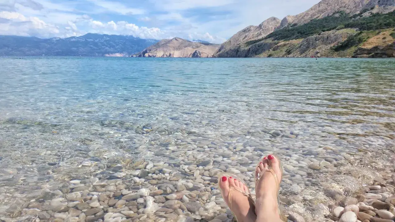Stopy w klapkach na kamienistej plaży, krystalicznie czysta woda i góry w tle. Idealny dzień na wyspie Krk w Chorwacji.