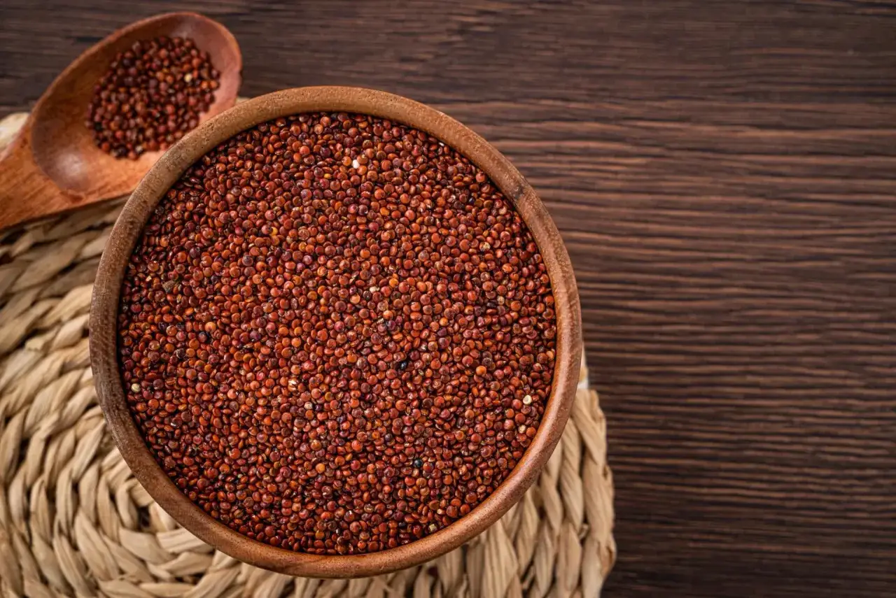 Una ciotola di legno piena di quinoa rossa, con un cucchiaio di legno accanto, su un sottobicchiere intrecciato.
