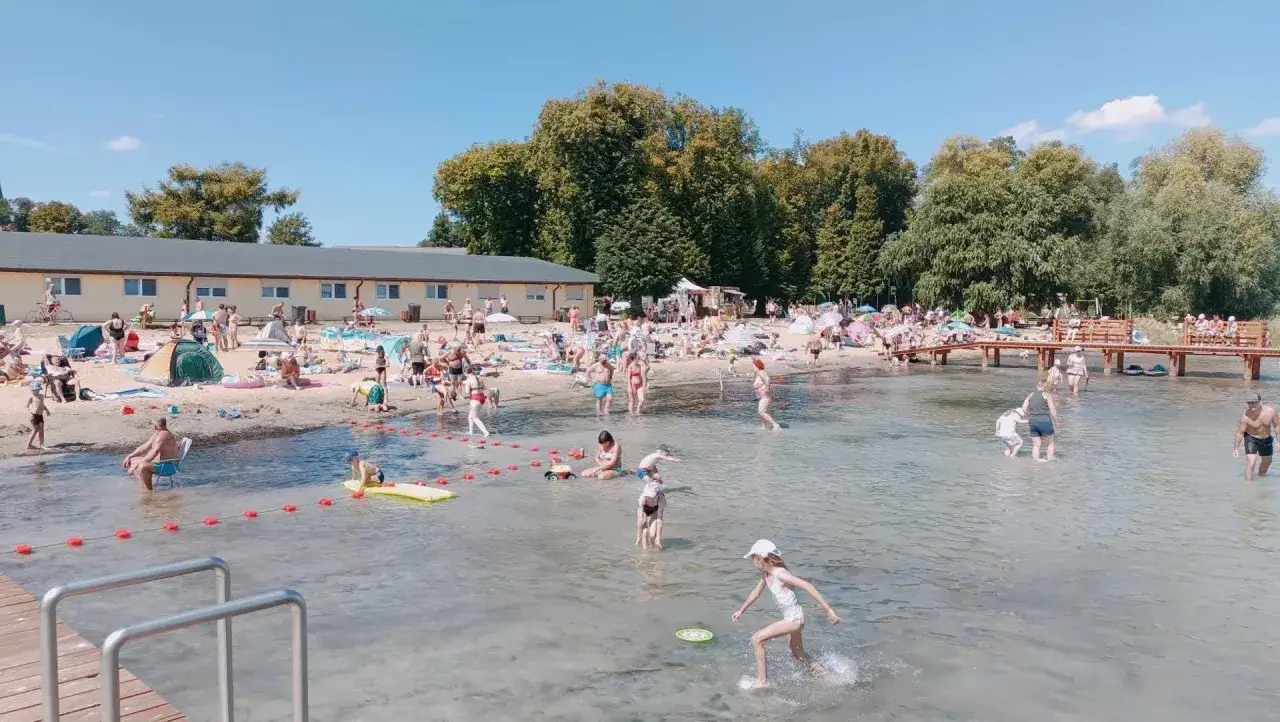 Słoneczna plaża pełna ludzi kąpiących się i opalających. Dzieci bawią się w wodzie, a dorośli odpoczywają na piasku.