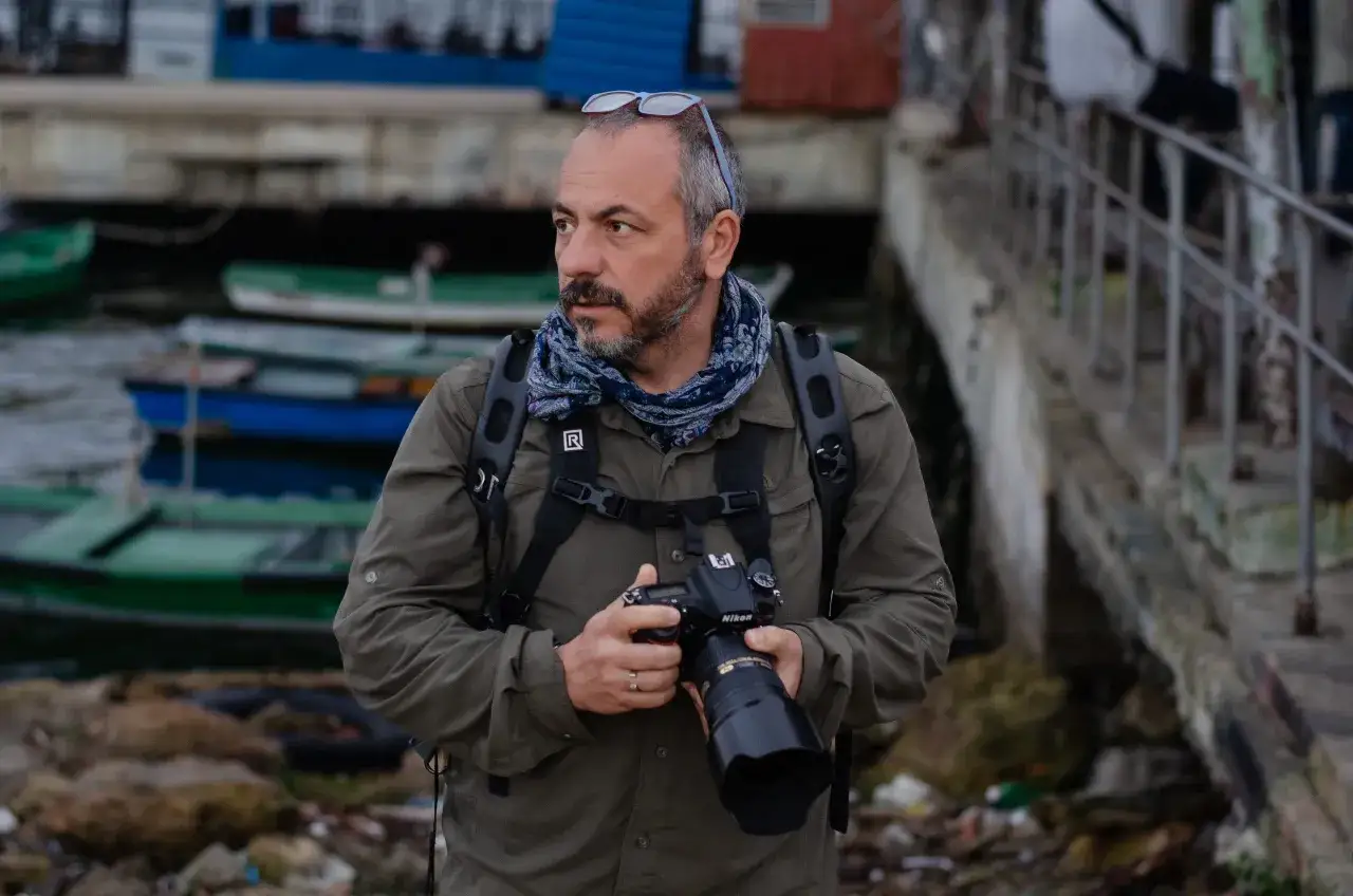 Un uomo con macchina fotografica, occhiali da sole sulla testa e sciarpa, pronto a documentare la realtà. Il suo sguardo è attento, tipico di un fotoreporter.