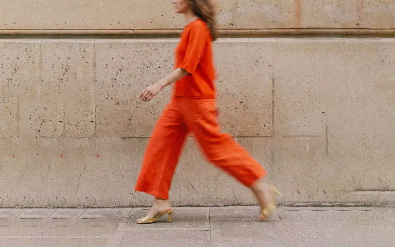 Frau in leuchtend orangefarbenem Leinen-Outfit im Sommer, das sich vor einer Steinwand bewegt.