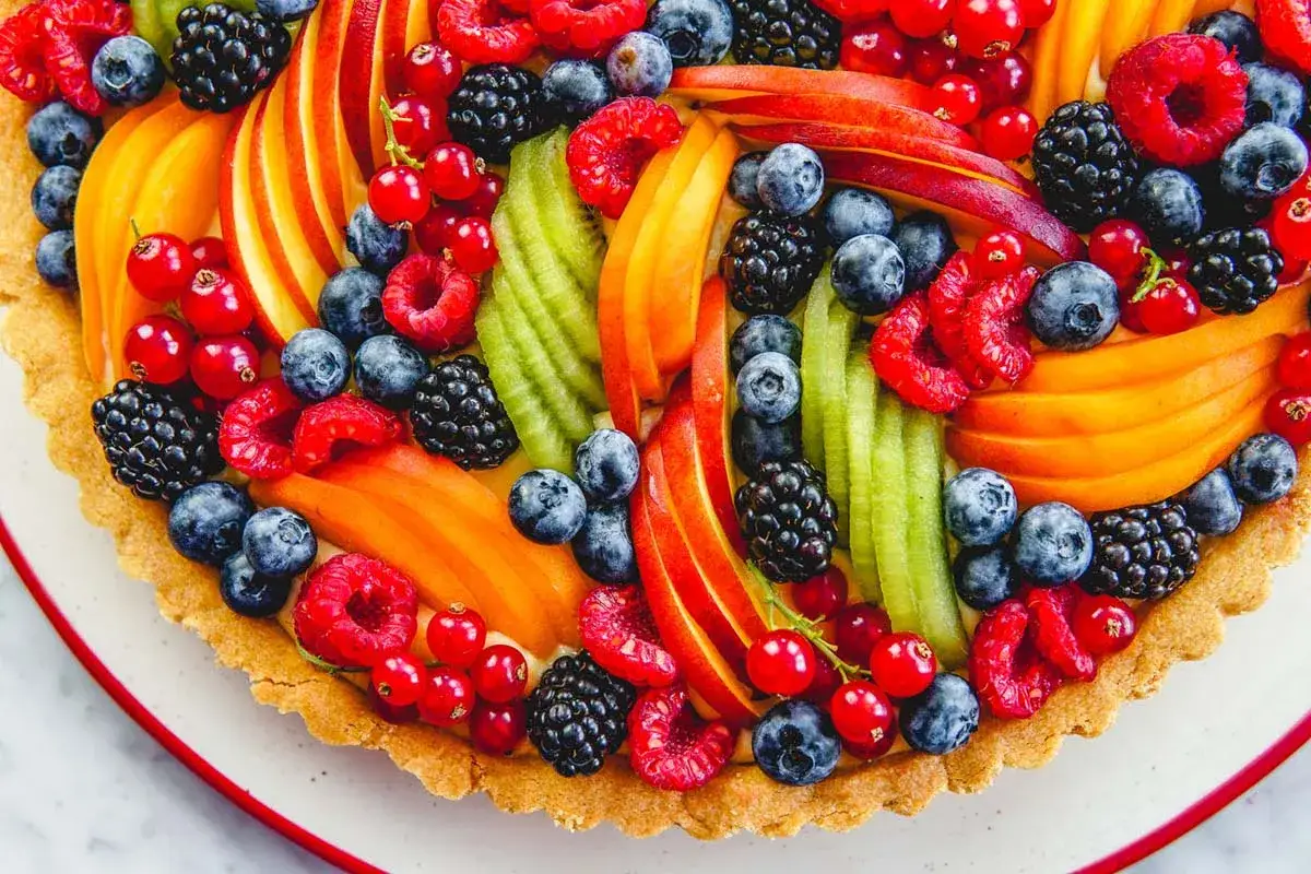 Crostata di frutta con fragole, mirtilli, lamponi, ribes, pesche e kiwi. Le decorazioni sono disposte a raggiera, creando un effetto visivo vibrante e invitante.