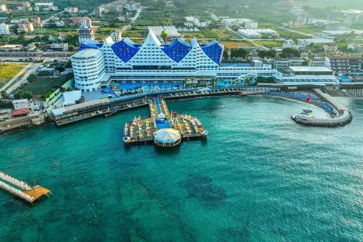 Widok na hotel w kształcie górskich szczytów w Orange County Alanya, z pomostem i turkusową wodą.