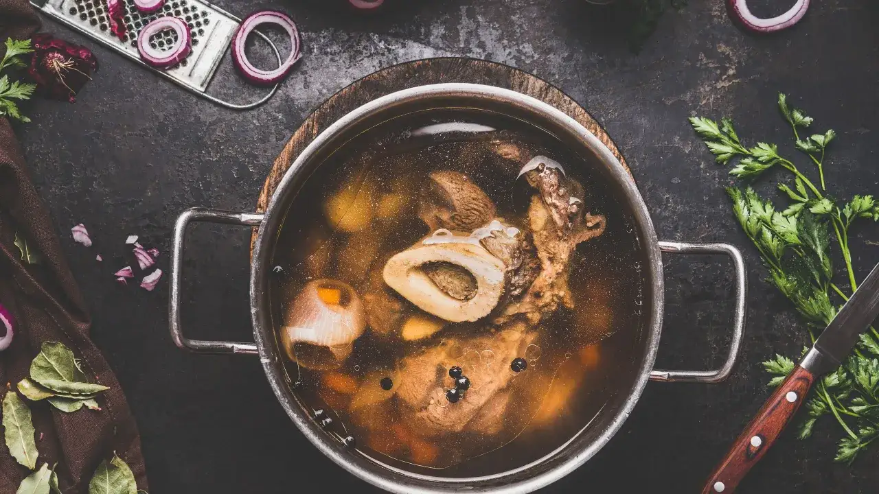 Un pentolone pieno di brodo di carne fumante, con ossa, verdure e grani di pepe. Ingredienti freschi come cipolla e prezzemolo completano la scena.