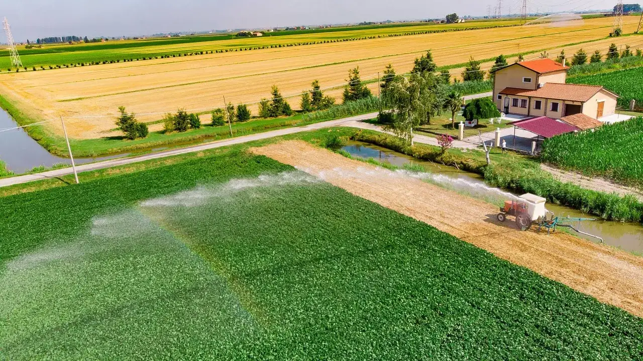 Un trattore irriga un campo verde, creando un arcobaleno. La scena mostra la moderna resa agricola con campi dorati e mais sullo sfondo.