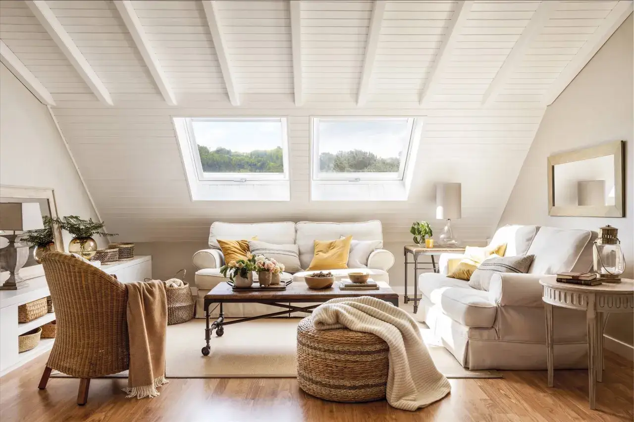 Ideas para decorar un ático: sala de estar luminosa con sofás blancos, mesa de centro de madera, sillón de mimbre y ventanas en el techo.