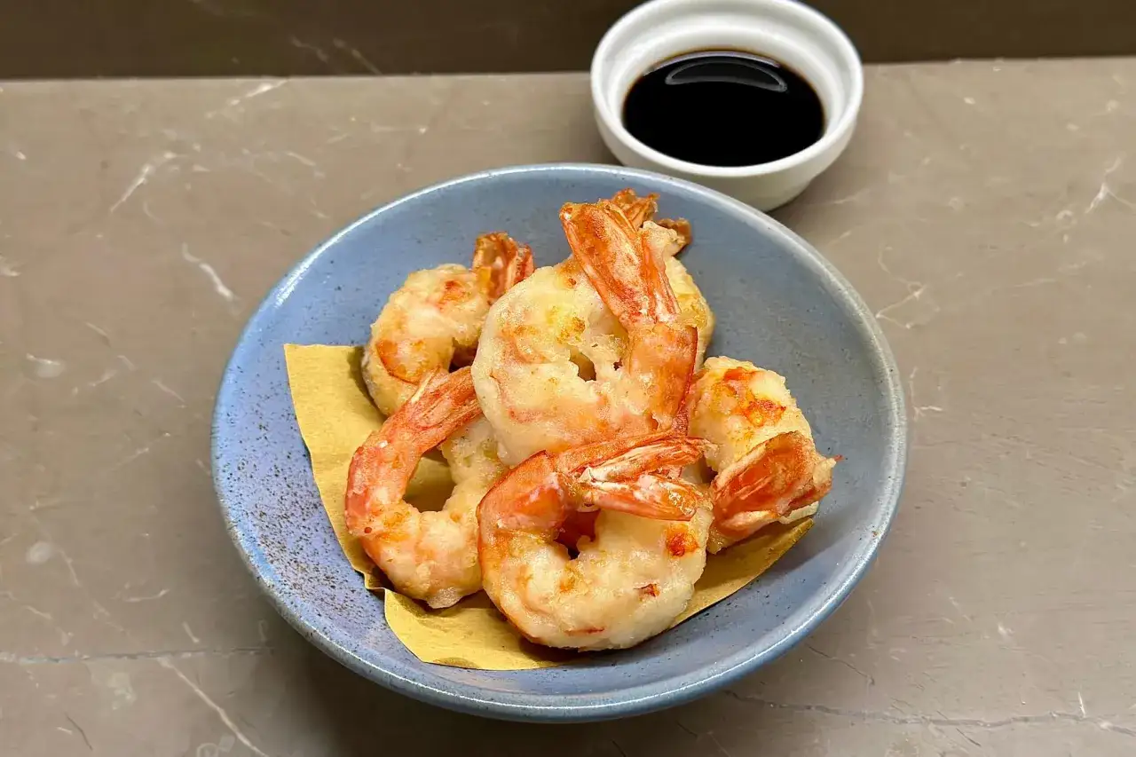 Gamberi in tempura dorati e croccanti serviti in una ciotola blu, con una piccola ciotola di salsa scura sullo sfondo.