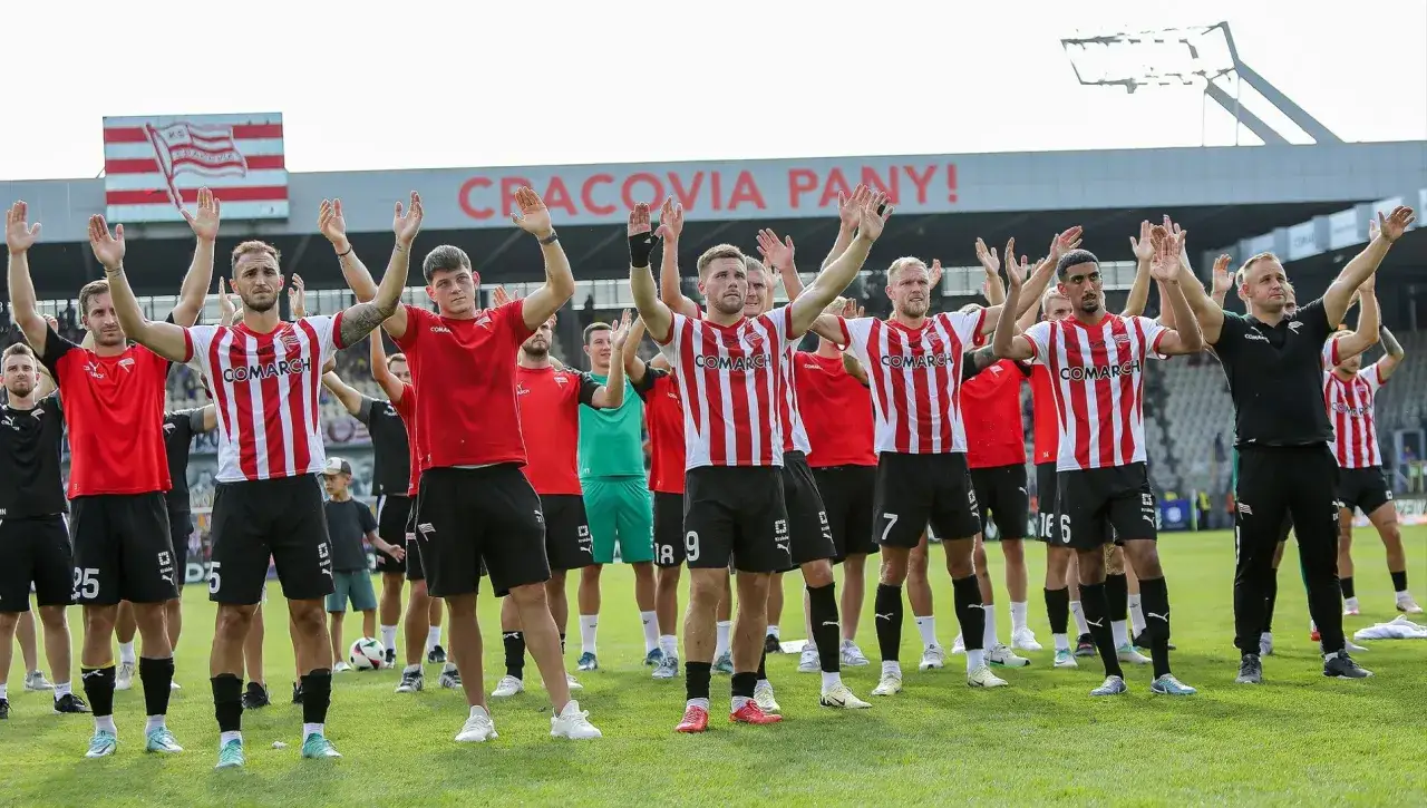 Piłkarze Cracovii w pasiaste koszulki pozdrawiają kibiców. W tle napis 