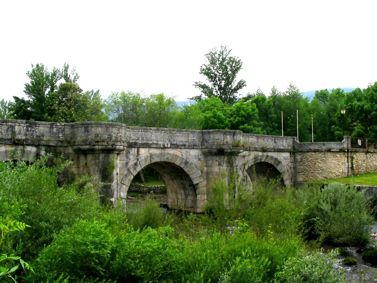 Puente de piedra con varios arcos en Rascafr&iacute;a, Madrid, rodeado de vegetaci&oacute;n exuberante y &aacute;rboles frondosos.