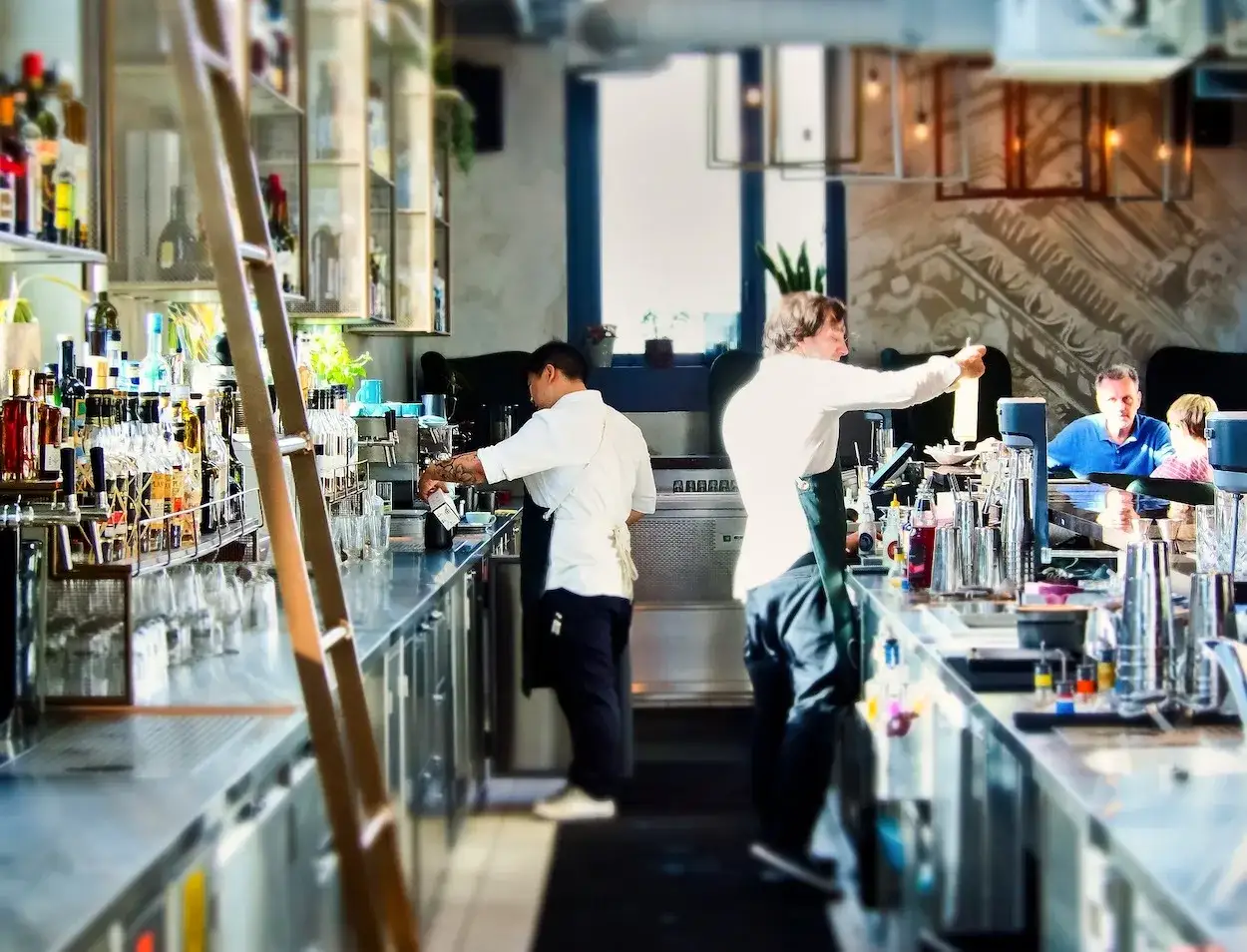 Barra de bar con camareros preparando bebidas. Un ejemplo de que es hosteler&iacute;a en Espa&ntilde;a, con clientes disfrutando en el fondo.