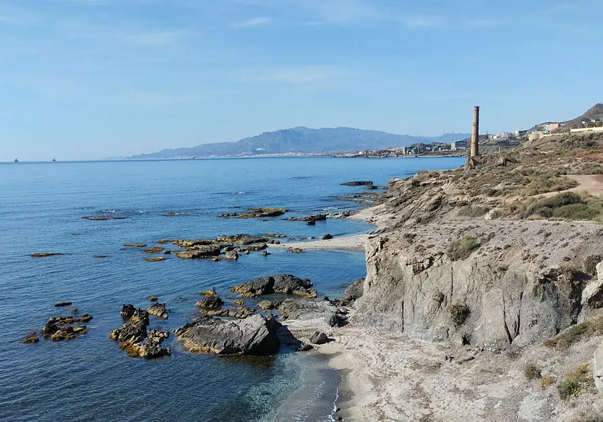 Costa rocosa con calas de Villaricos, mar azul y una chimenea industrial al fondo.