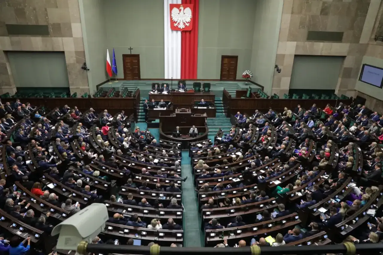 Posiedzenie Sejmu, gdzie trwa **obstrukcja** parlamentarna. Widać wielu posłów siedzących w ławach, flagi Polski i UE, a także godło państwowe.
