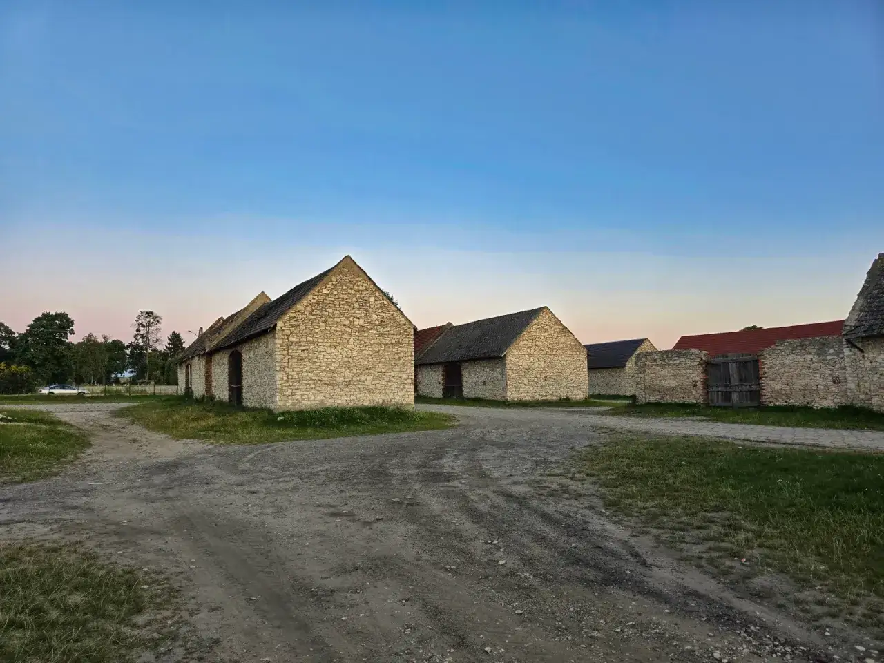 Kamienne stodoły w Żarkach, oświetlone łagodnym światłem zachodzącego słońca, stoją w rzędzie przy piaszczystej drodze.