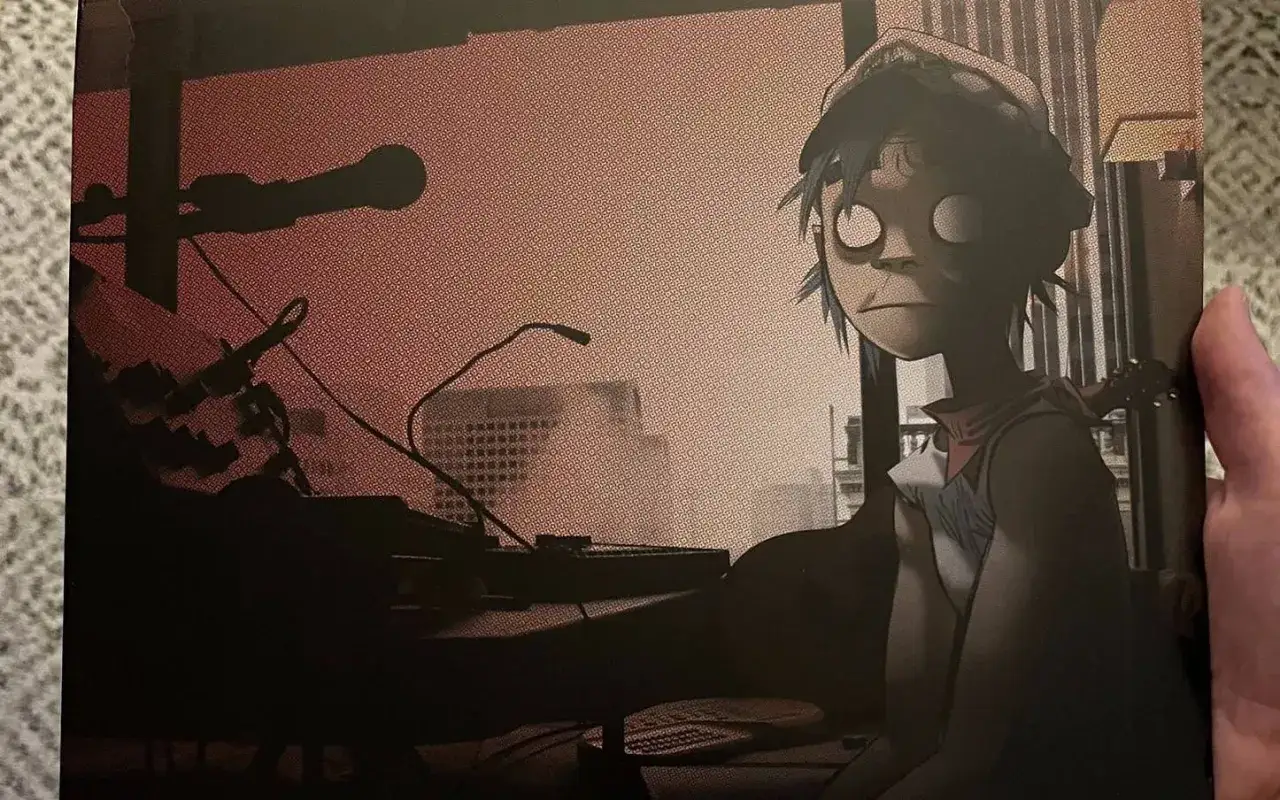 Okładka albumu Gorillaz The Fall. 2-D siedzi przy konsoli, otoczony sprzętem studyjnym i miejskim krajobrazem.