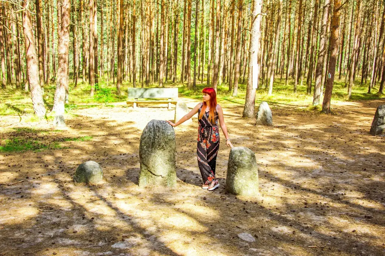 Kobieta z rudymi włosami stoi przy kamieniach. W tle las i ławka. To tajemnicza stanica nad kamiennymi kręgami.