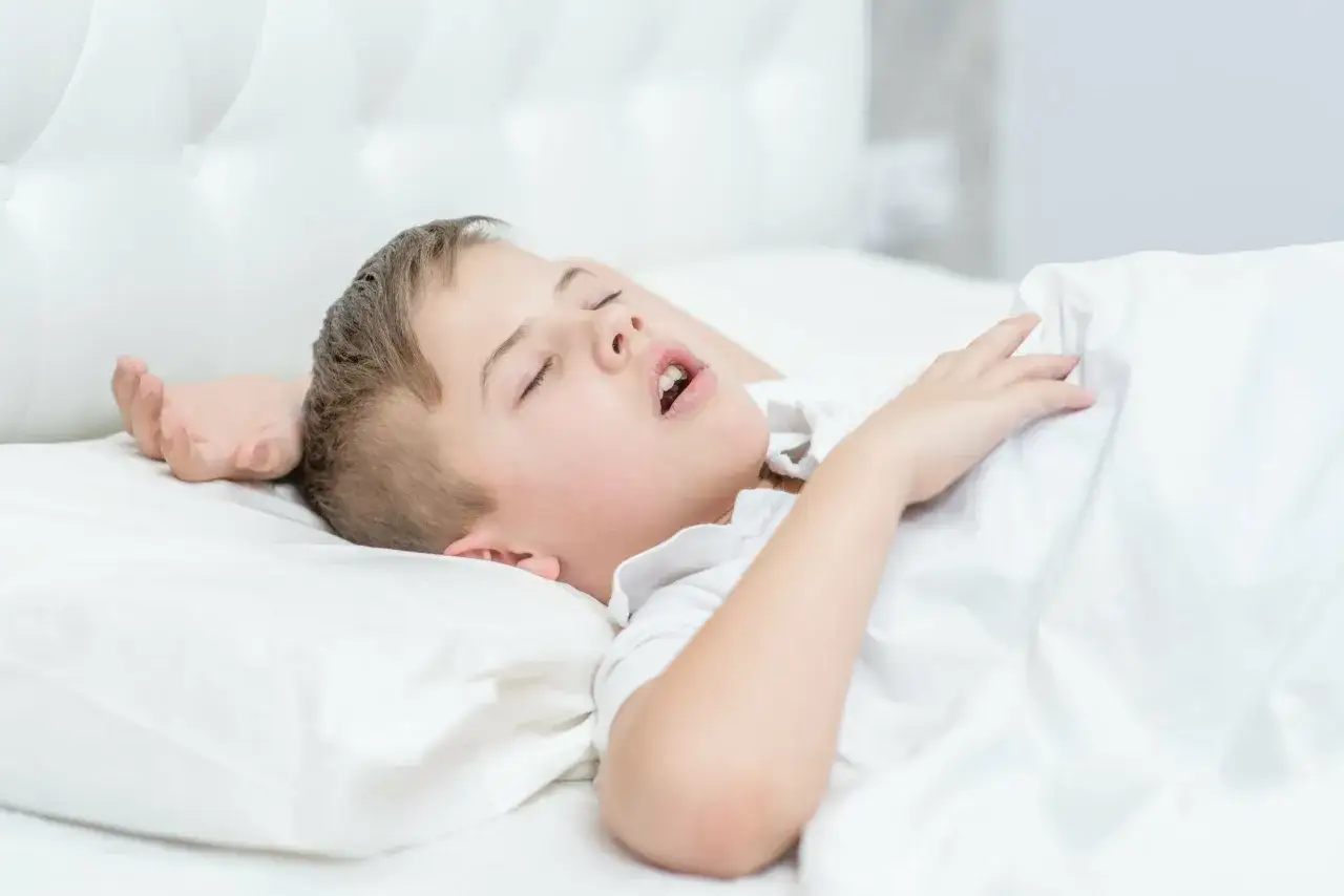 Chłopiec śpi z otwartą buzią, wtulony w białą pościel. Jego ręka spoczywa na kołdrze.