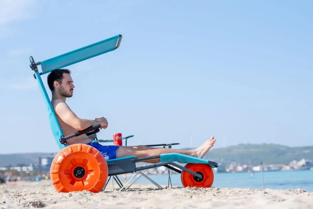 Uomo rilassato su una sedia job con ruote arancioni sulla sabbia, con il mare e il cielo sullo sfondo.