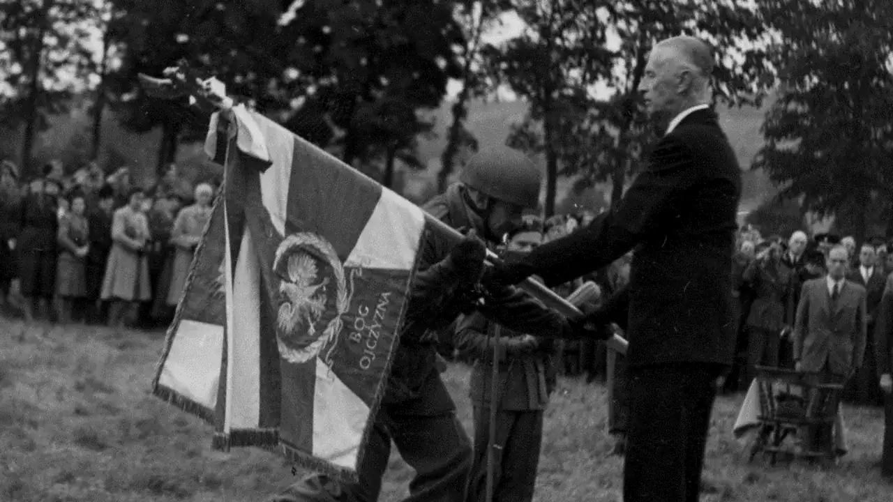 Uroczystość przekazania sztandaru z hasłem Bóg Honor Ojczyzna. Mężczyzna w garniturze odbiera go od żołnierza.