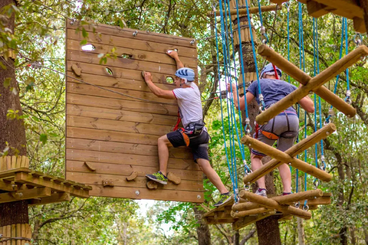 Ragazzi si divertono in un parco avventura Puglia, arrampicandosi su una parete di legno e attraversando un ponte sospeso tra gli alberi.