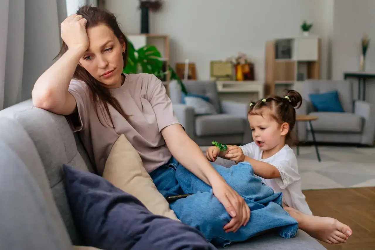 Zmęczona mama zamyślona, podczas gdy dziecko bawi się obok. Czasem nawet spokojne chwile przy dziecku mogą przypominać kłótnie.