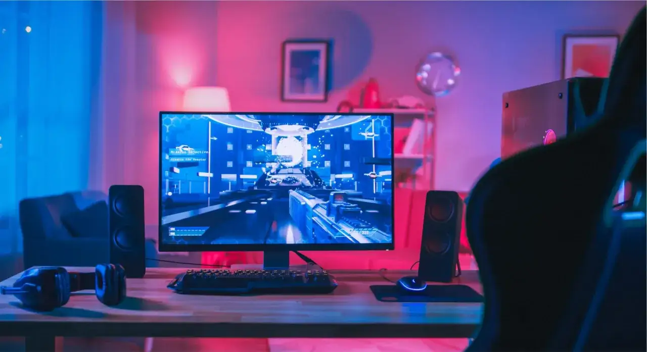 Un bureau de gamer avec un écran affichant un jeu fps pc, des enceintes, un casque et un fauteuil de jeu, le tout baigné de lumières bleues et roses.