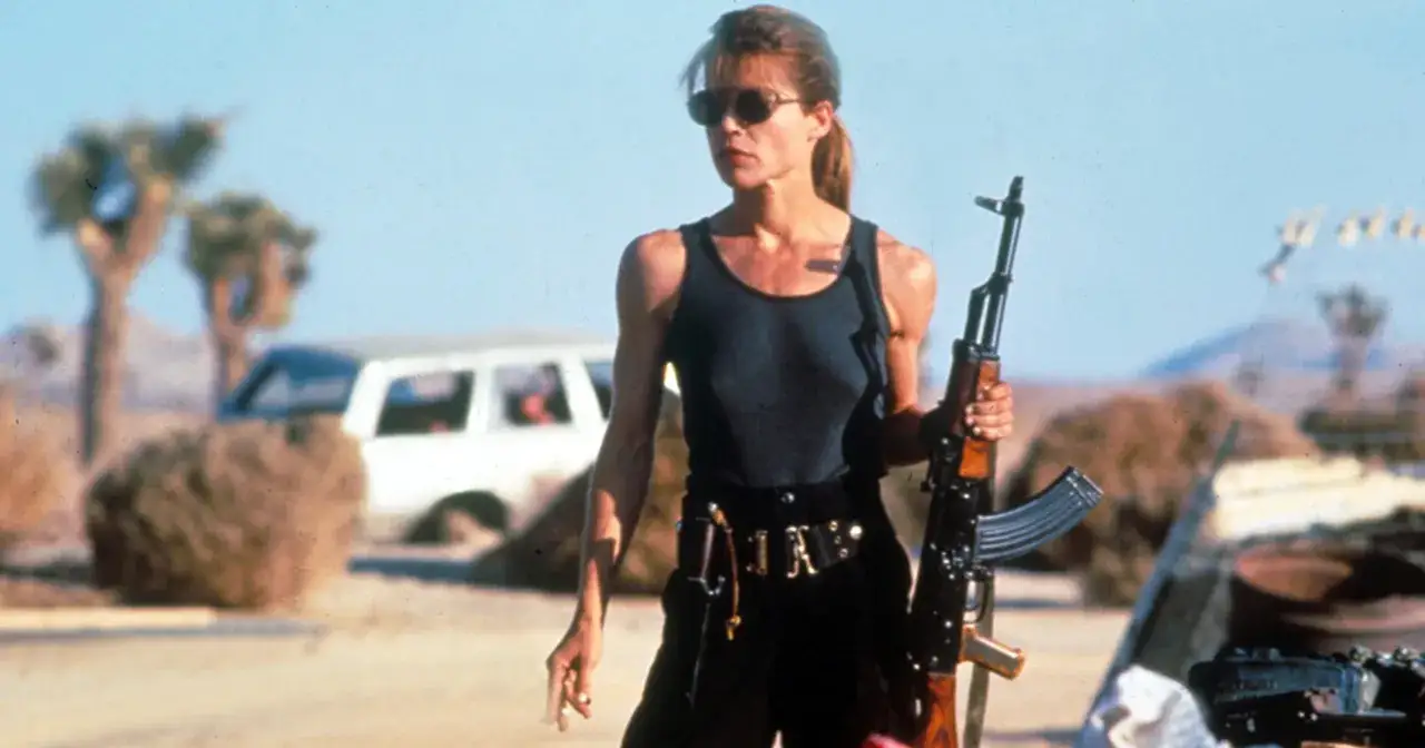 Linda Hamilton w roli Sarah Connor, gotowa do walki z karabinem w postapokaliptycznym krajobrazie. Ikona kina akcji, znana z wielu filmów i seriali.