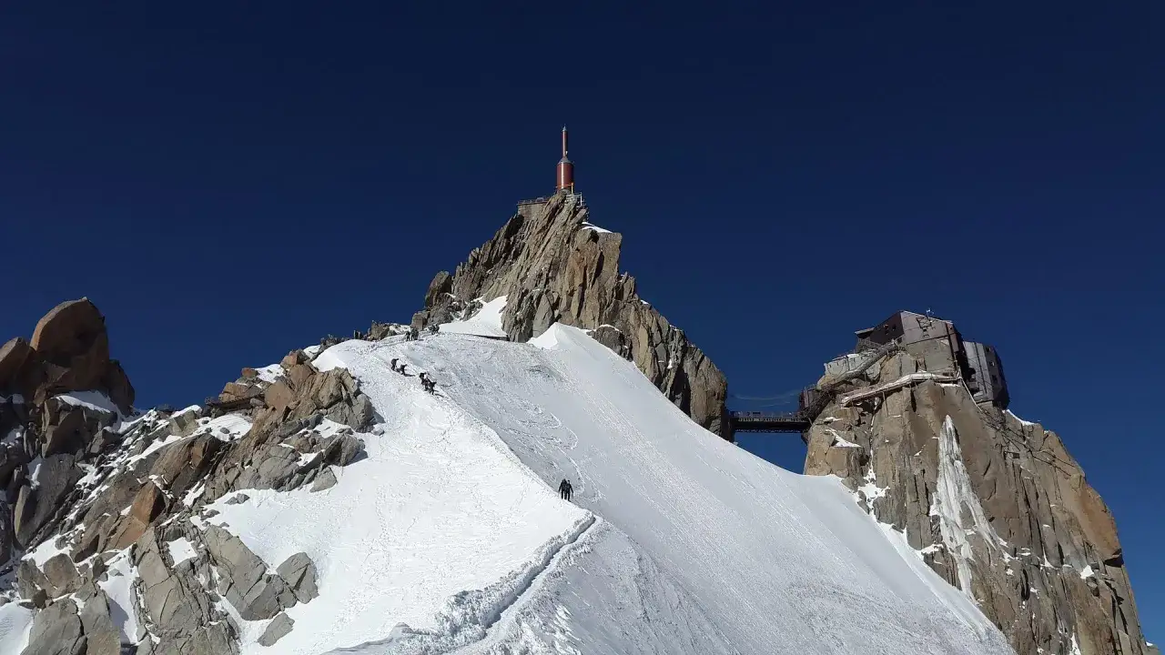 Wspinacze na ośnieżonym stoku, zbliżający się do szczytu z wieżą przekaźnikową. Widok na masyw **Mont Blanc**.