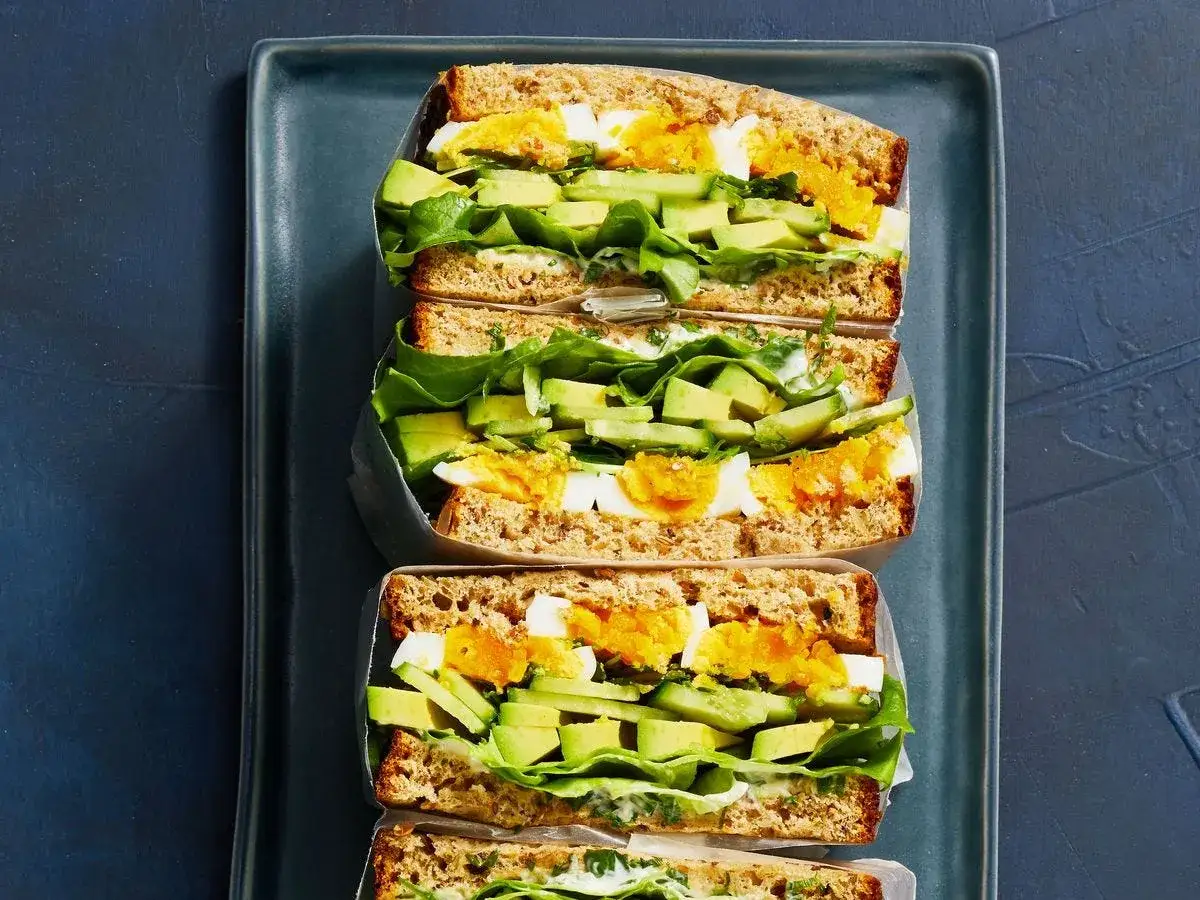 Tres sándwiches saludables apilados en un plato azul oscuro. Rellenos de aguacate, huevo duro, pepino y lechuga.