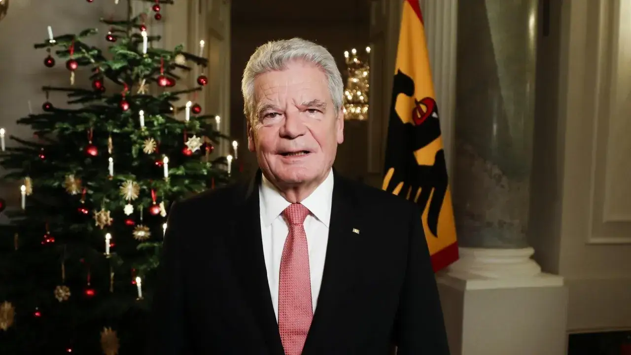 Ein Mann im Anzug steht vor einem Weihnachtsbaum und der deutschen Flagge. Er h&auml;lt eine Rede, die als beste Weihnachtsrede gilt.
