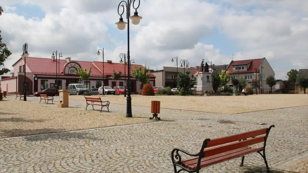 Rynek w Ślesinie z pomnikiem, ławkami i budynkami. Atrakcje tego miejsca zachęcają do spacerów.