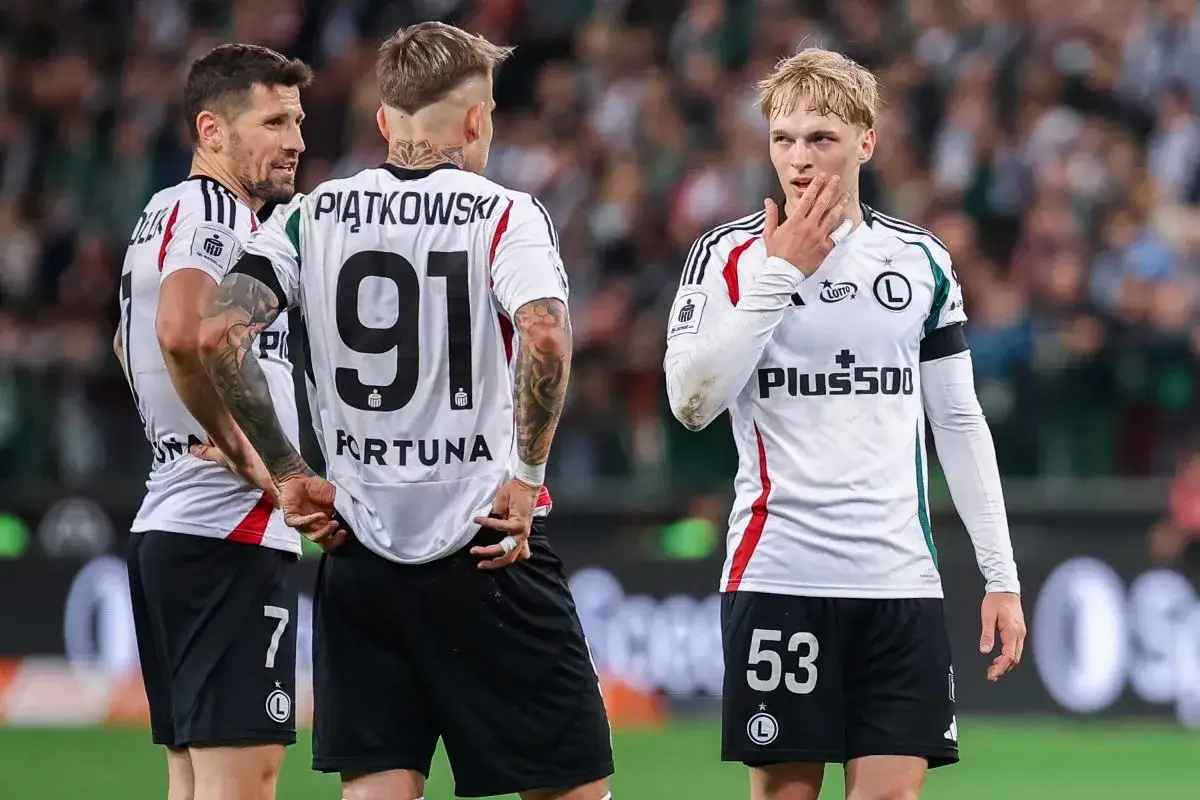 Trzech zawodników Legii Warszawa w białych strojach dyskutuje na boisku. Na koszulkach numery 91, 7 i 53.