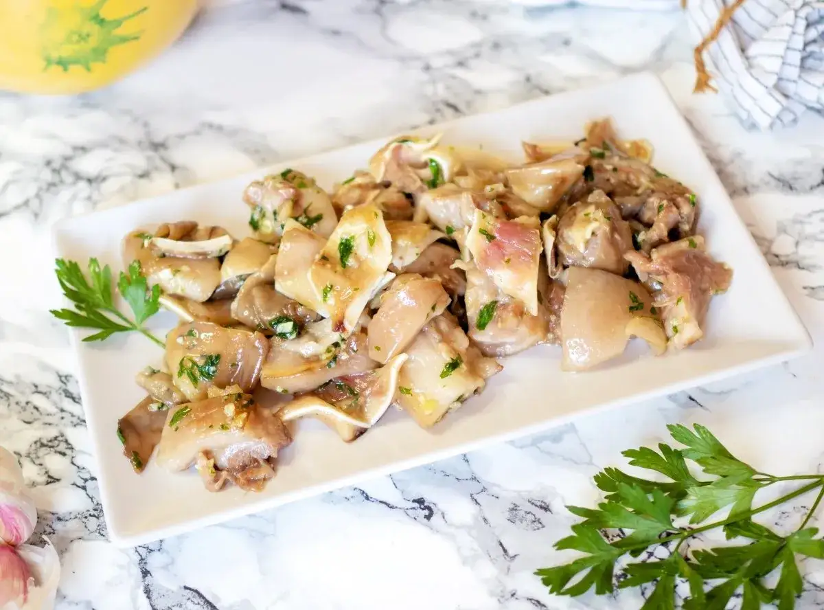 Oreja de cerdo marinada con ajo y perejil, servida en un plato blanco sobre una superficie de m&aacute;rmol.