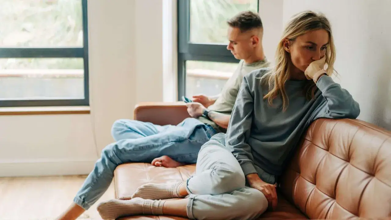 Coppia sul divano, lei pensierosa, lui al telefono. Un momento di distanza, dove si può **non sentirsi capiti dal partner**.
