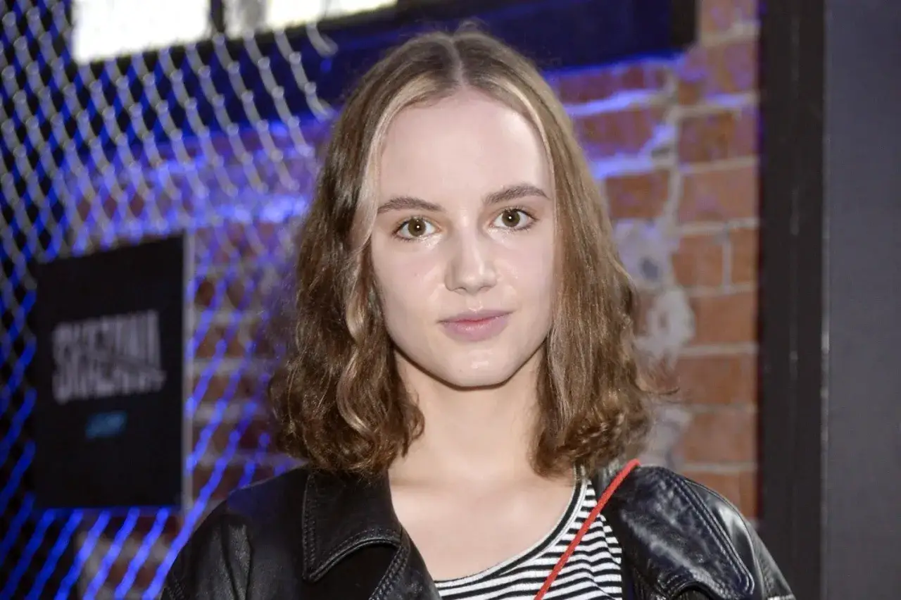 Martyna Byczkowska w czarnej ramonesce i bluzce w paski, z rudym akcentem na włosach, pozuje na tle ceglanej ściany i niebieskiej siatki.