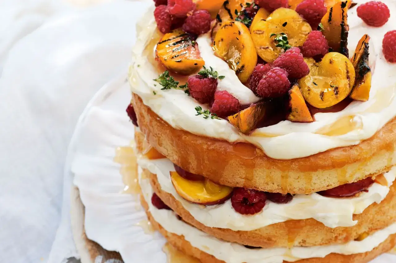 Puszysty tort z bitą śmietaną i mascarpone, przełożony owocami i udekorowany malinami oraz grillowaną brzoskwinią.